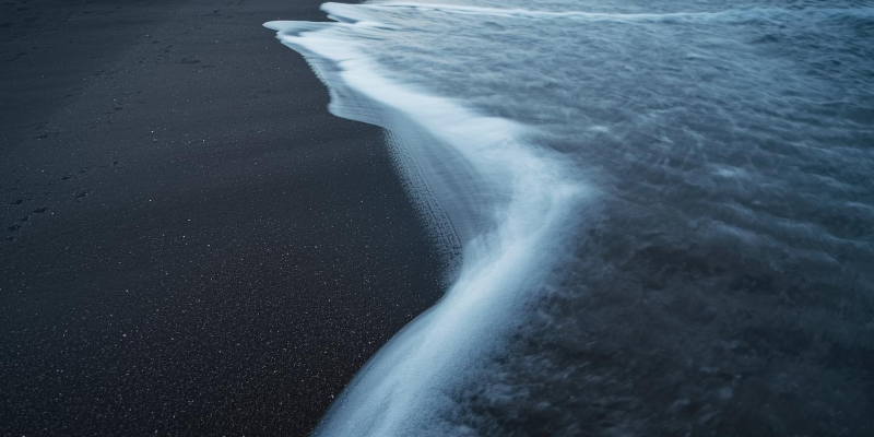 Dark sand beach with wave edge — the retention boundary where churn erodes growth